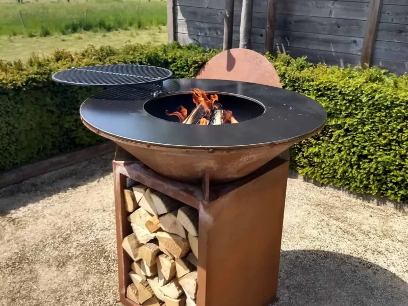 Cortenstaal BBQ met draaibaar grillrooster boven het vuur, windscherm en een onderstel vol brandhout