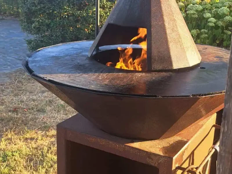 Cortenstaal BBQ op maat met rookkap en schoorsteen boven de vuurschaal en een open houtopslag