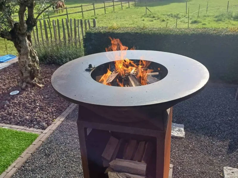 Brandende cortenstaal BBQ met brede bakplaat rond de vuurschaal en houtopslag, in een tuin met zicht op de weide
